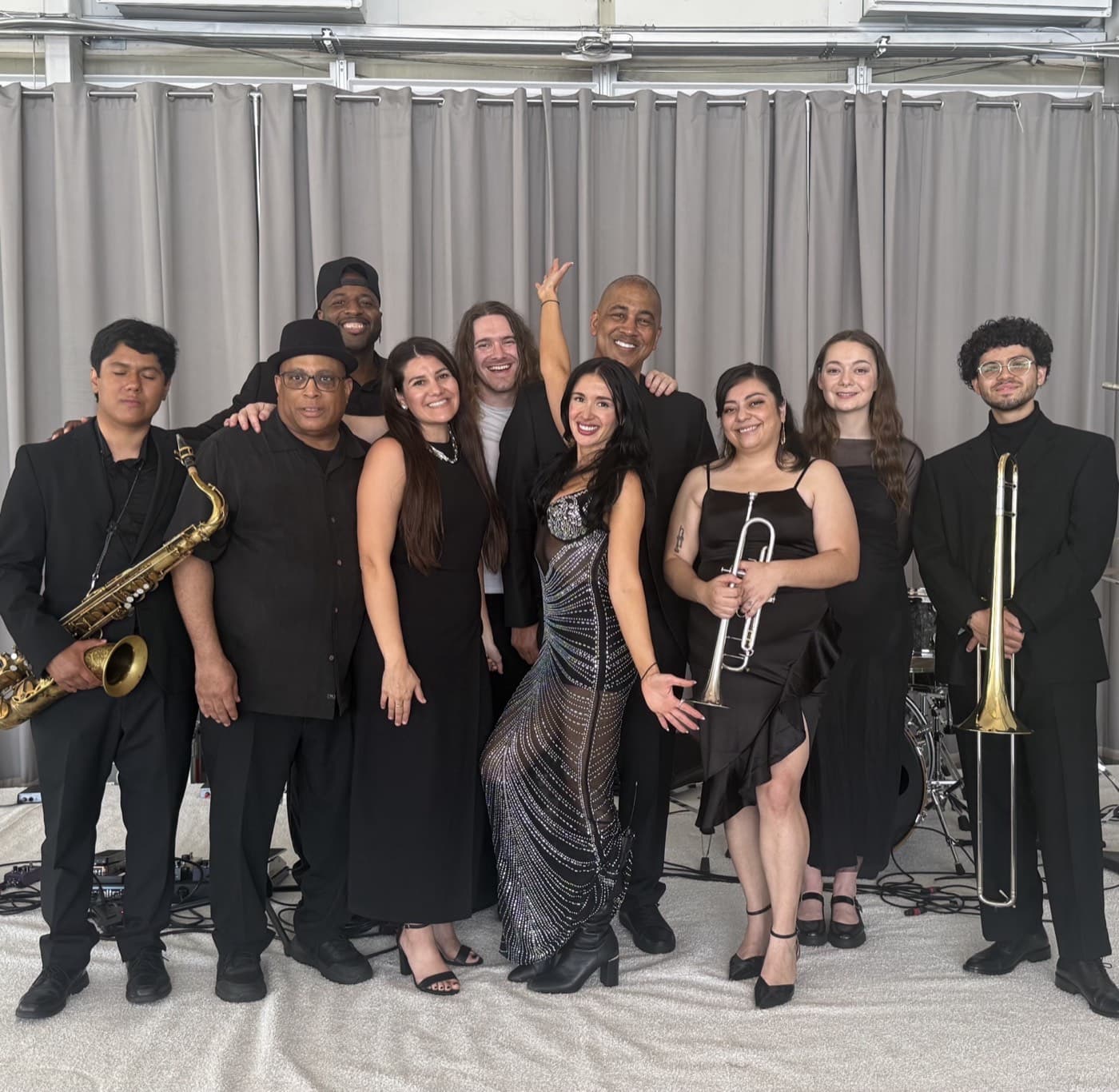 Jackie J & The Diamonds — full 9-piece band lineup in all black with horns, drums, and Jackie centered in a metallic dress.