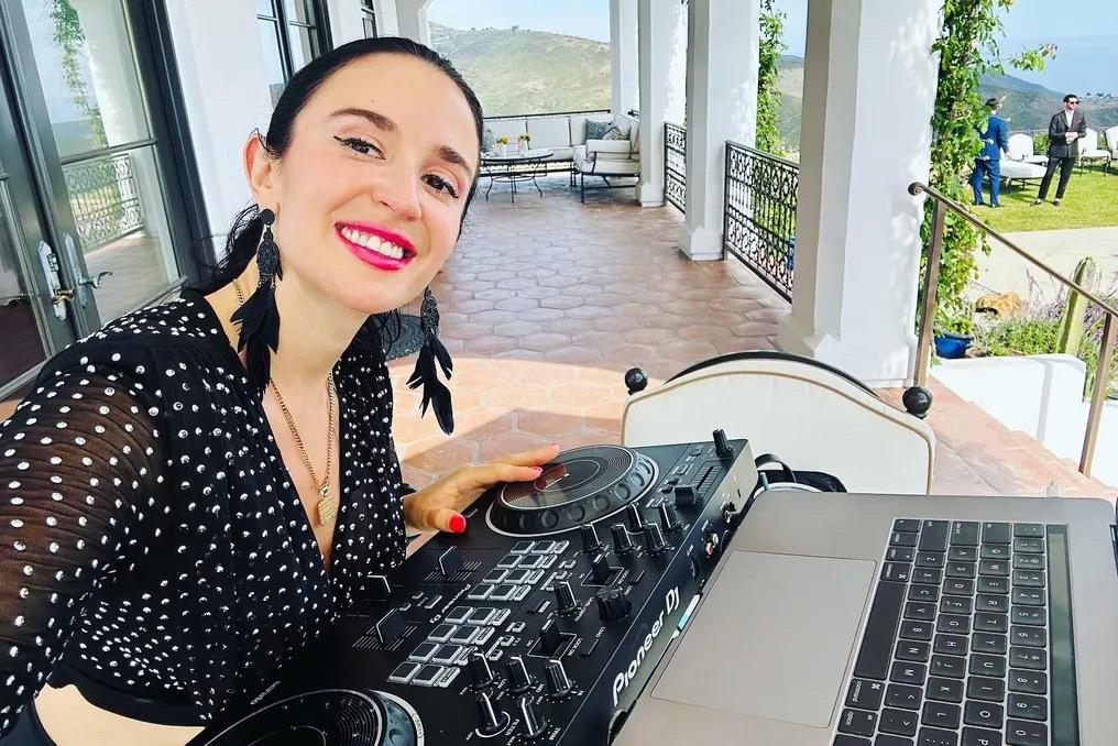 Jackie in a black-and-white polka dot dress smiling behind her DJ controller, hilltop villa in golden light.